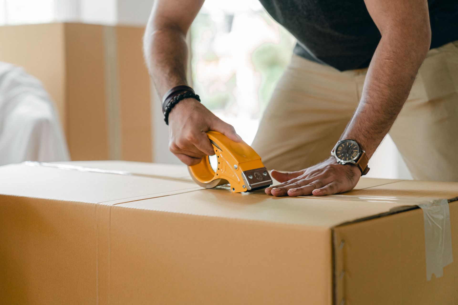 Crop unrecognizable young male in casual clothes packing cardboard box and sealing with scotch tape