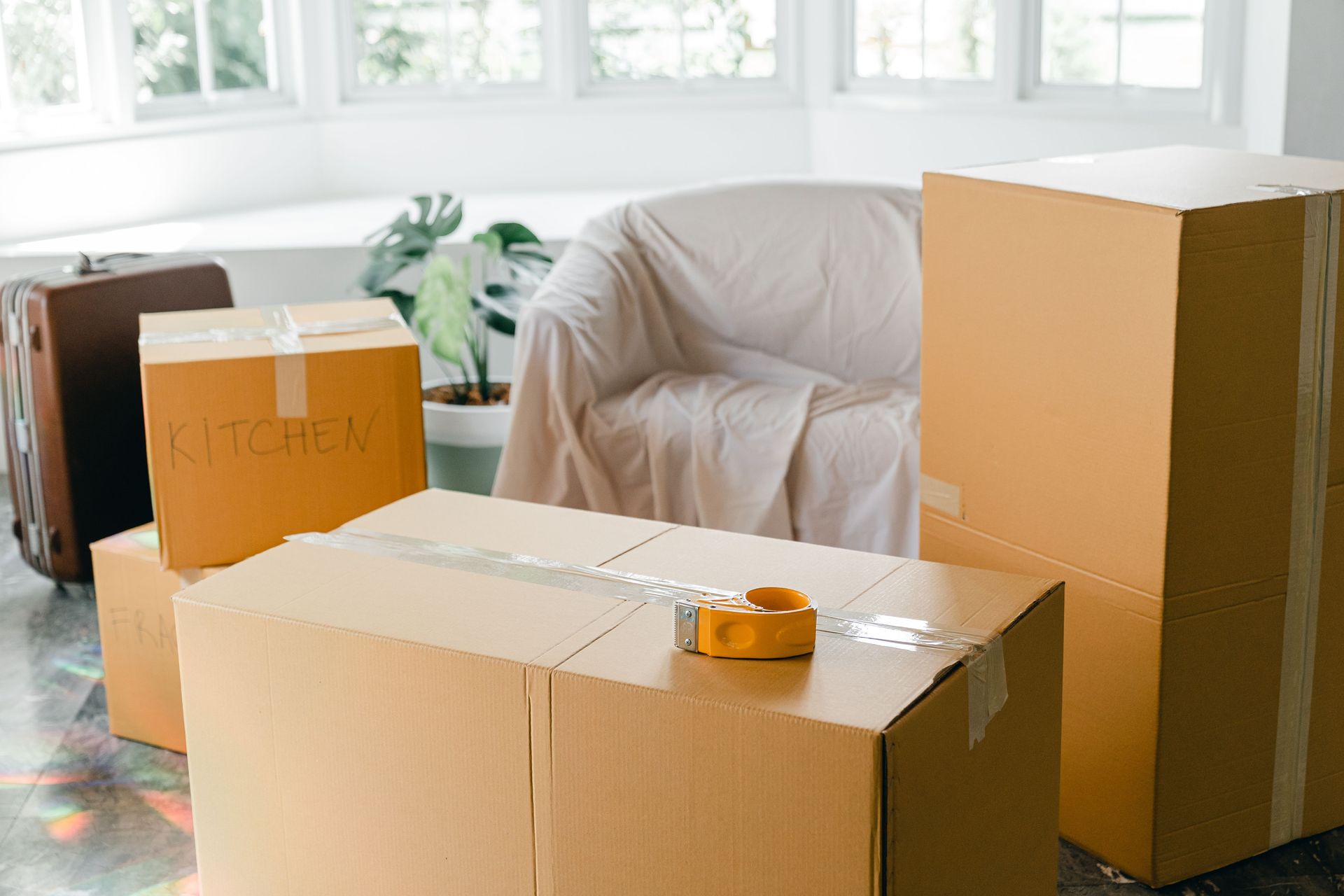 Various cardboard boxes with household belongings and suitcase placed on floor in bright room before relocation to new apartment